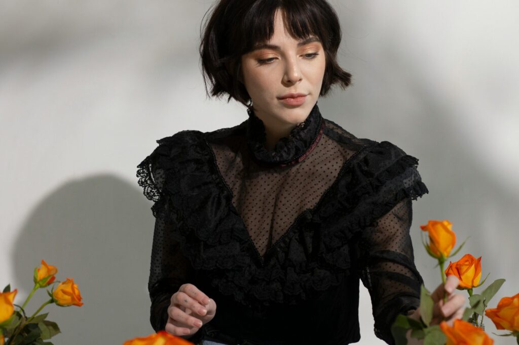 Elegant woman in stylish black blouse surrounded by orange roses in studio setting.