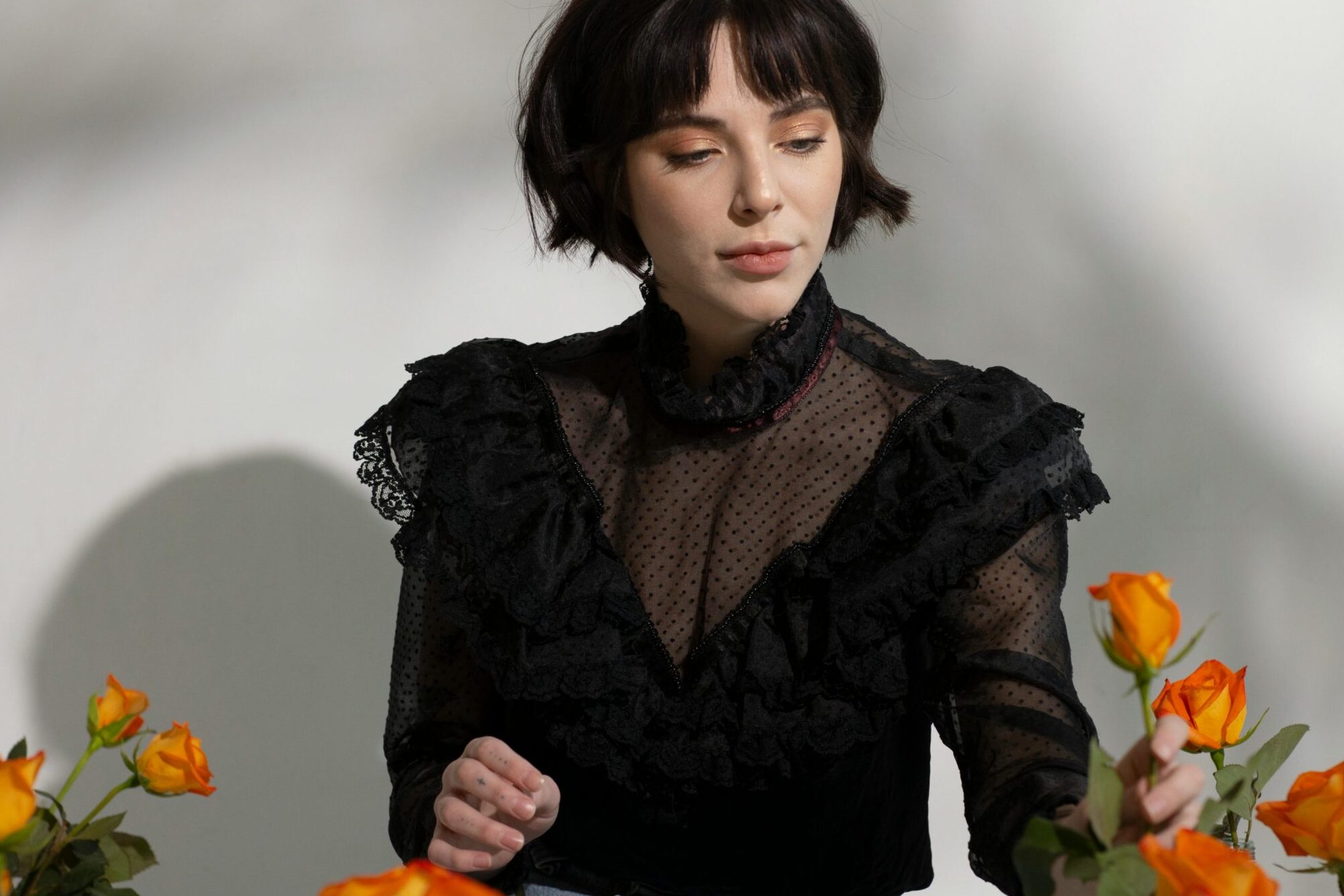 Elegant woman in stylish black blouse surrounded by orange roses in studio setting.